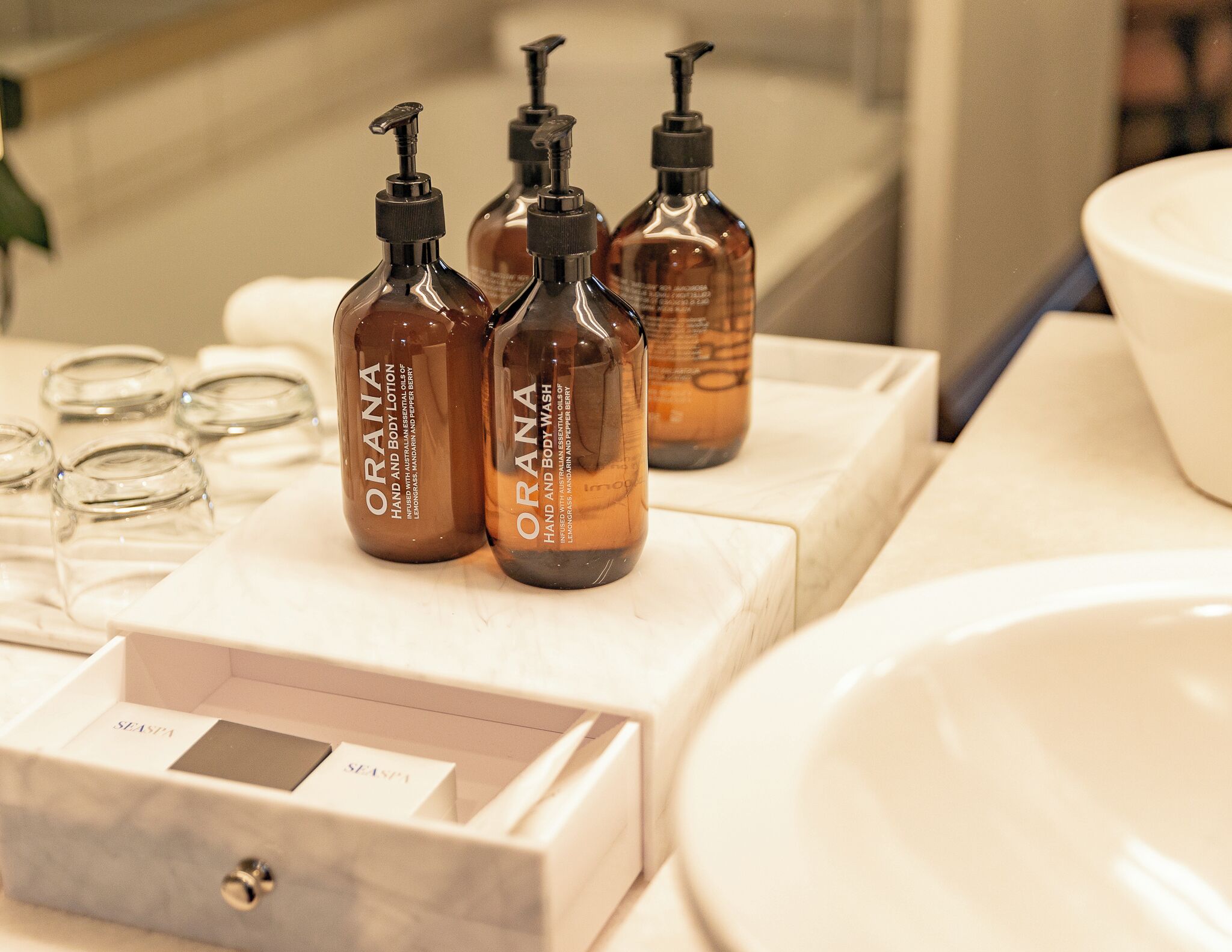 A bathroom with a marble sink, three bottles of ORANA hand and body wash, and a glass container.