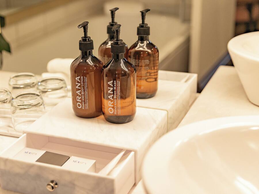A bathroom with a marble sink, three bottles of ORANA hand and body wash, and a glass container.
