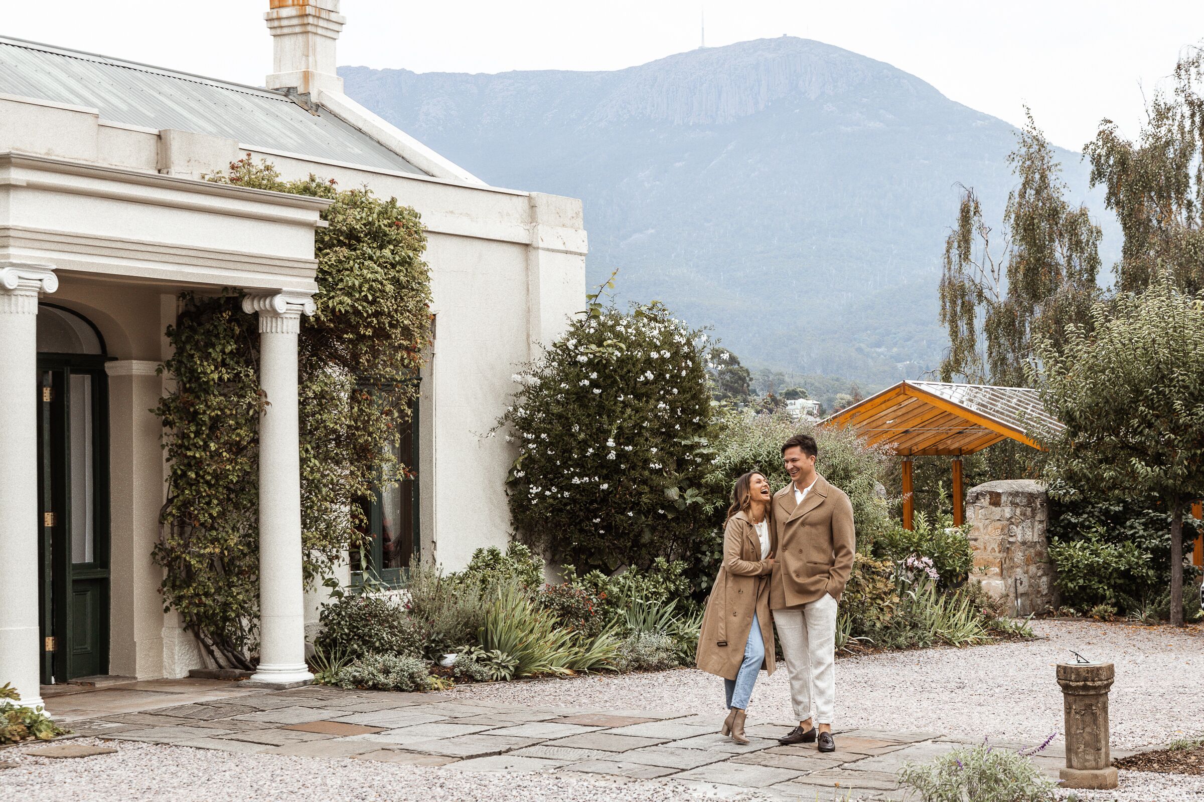 Una pareja está de pie en un jardín con un edificio blanco y montañas al fondo.