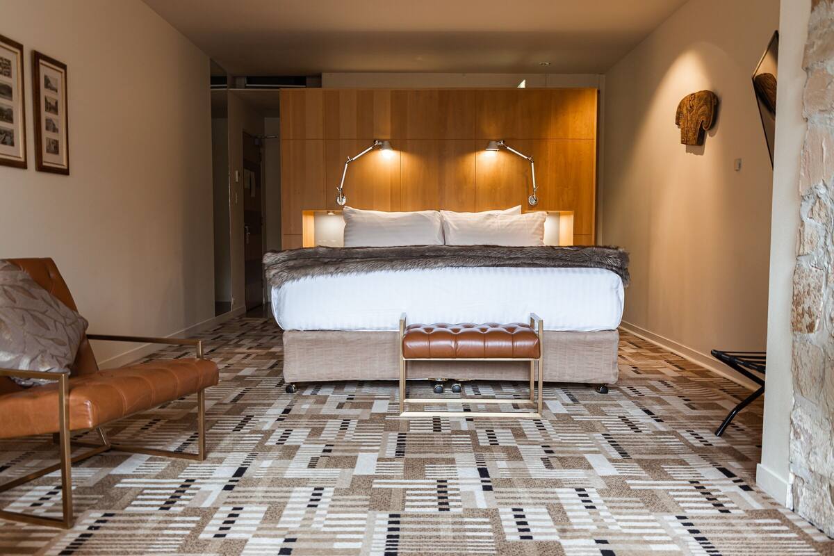 A bedroom with a large bed, two bedside lamps, a bench, a patterned carpet, and a wooden headboard.