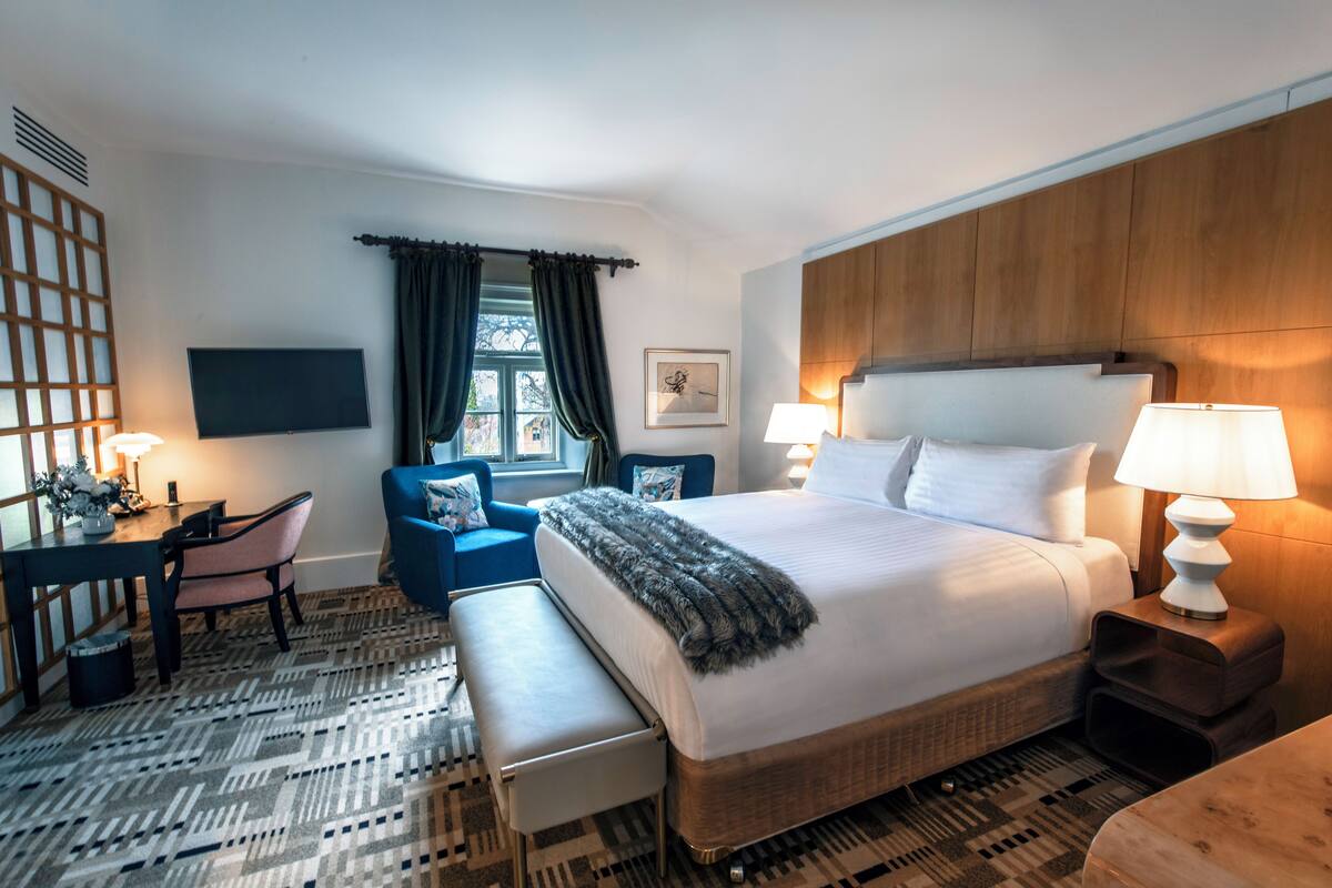 A hotel room with a large bed, bedside tables with lamps, a desk with a chair, a bench, and a view of the outdoors through a window with curtains.