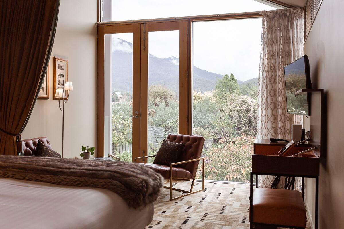 A bedroom with a large bed, a desk, a chair, and a view of mountains through a sliding glass door.