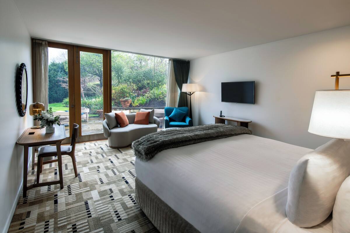 A hotel room with a large bed, a desk with a chair, a blue armchair, and a view of the garden through the sliding doors.