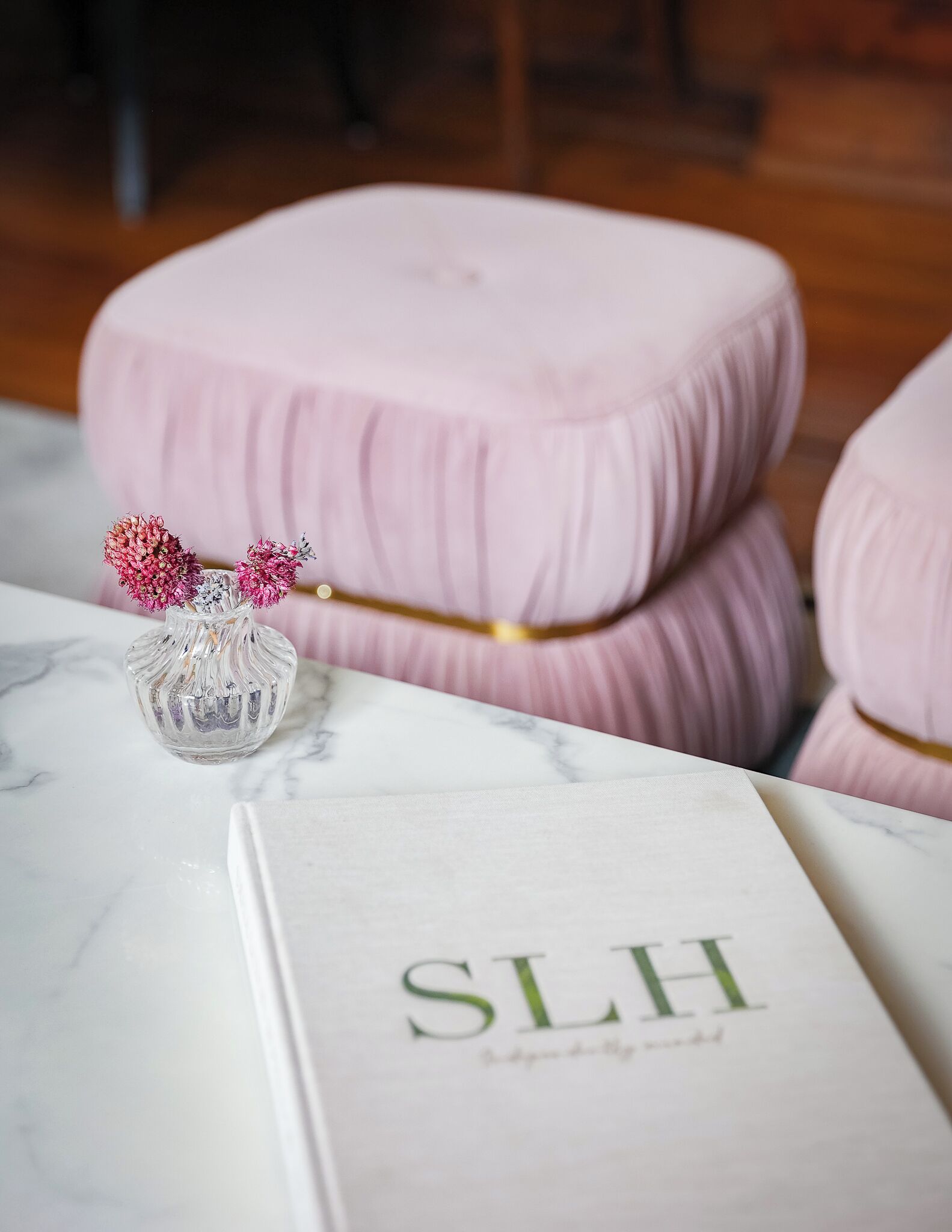 Ein Buch mit der Aufschrift 'SLH' auf einer Marmorplatte, eine transparente Glasvase mit getrockneten Blumen und zwei gepolsterte Hocker in Rosa.