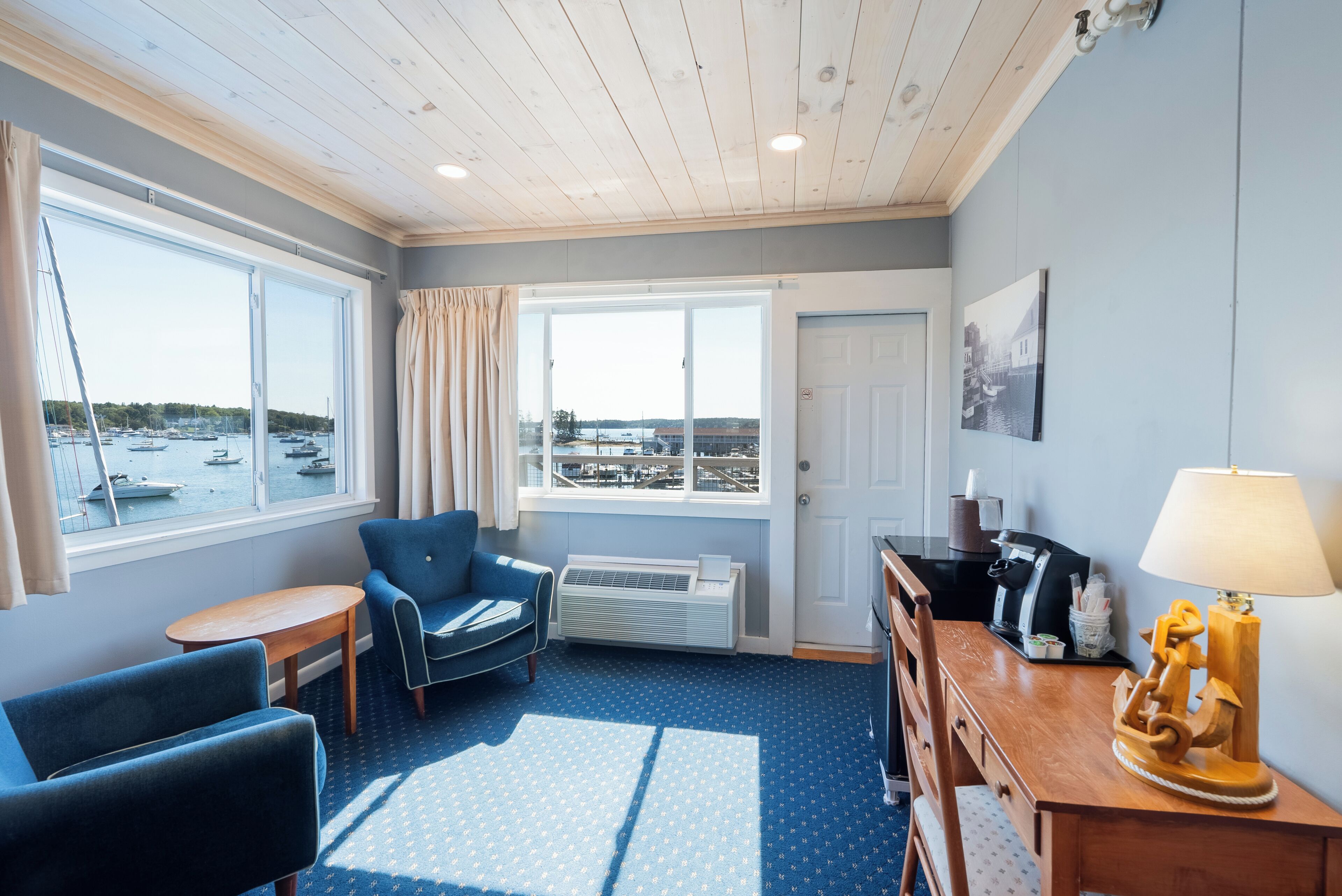 A room with a view of boats, a blue patterned carpet, a wooden desk, a blue armchair, and a lamp.