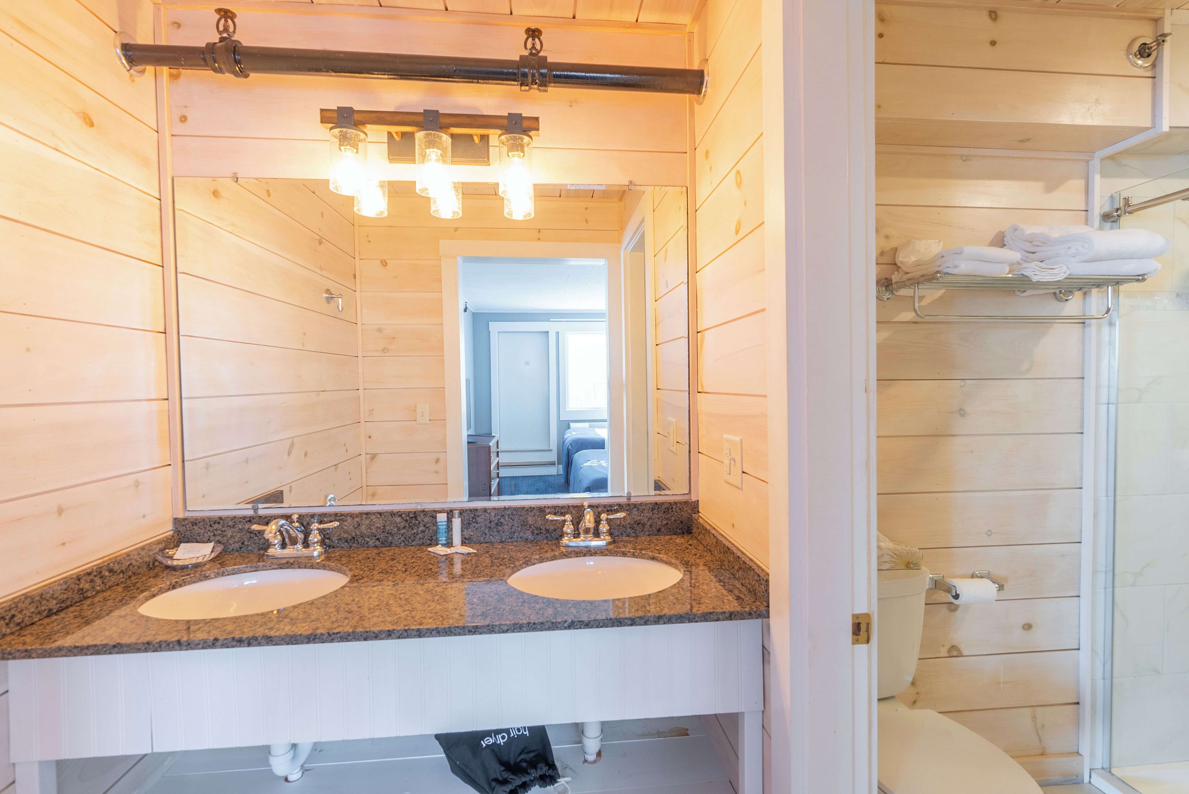 A bathroom with wooden walls, a granite countertop, two sinks, a mirror, and a shower area.