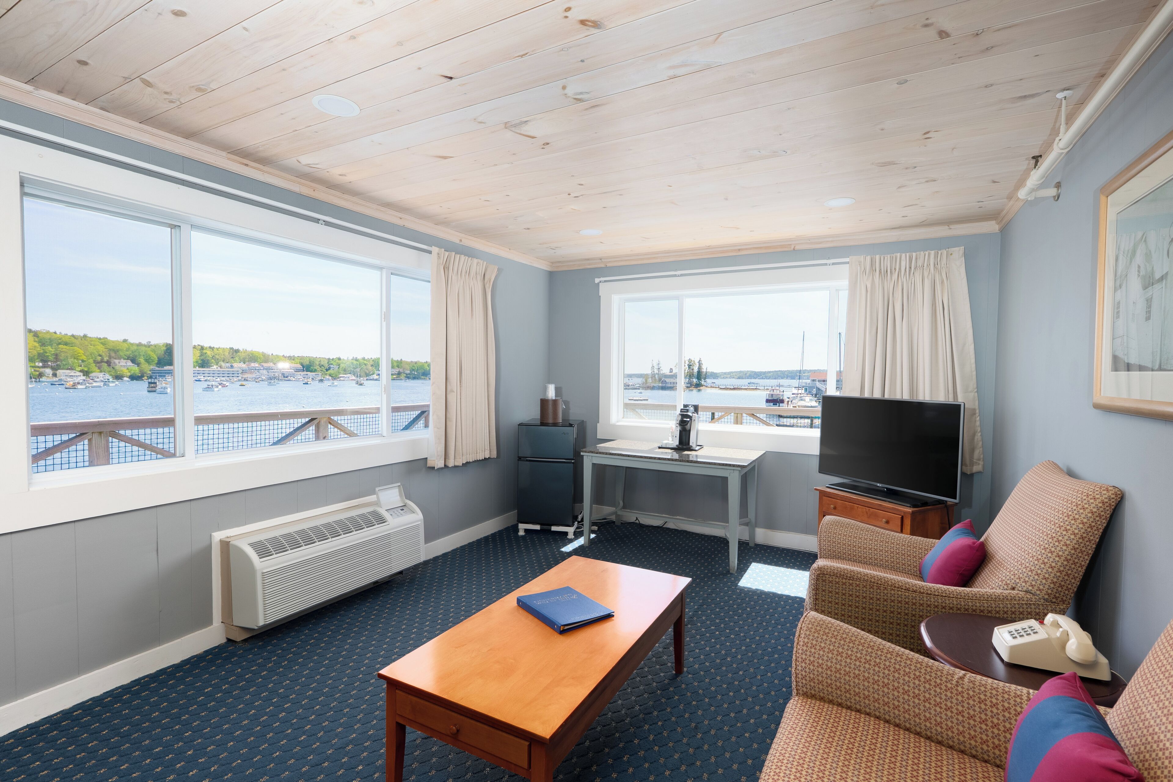 A room with a wooden ceiling, large windows with curtains, a desk, a television, a sofa, and a telephone.