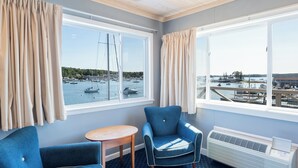 Ein Zimmer mit zwei blauen Sesseln, einem kleinen Holztisch und großen Fenstern mit Blick auf einen Yachthafen.