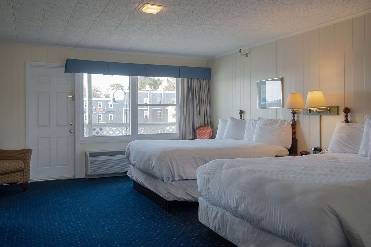 A hotel room with two beds, a window with curtains, a chair, and a lamp.