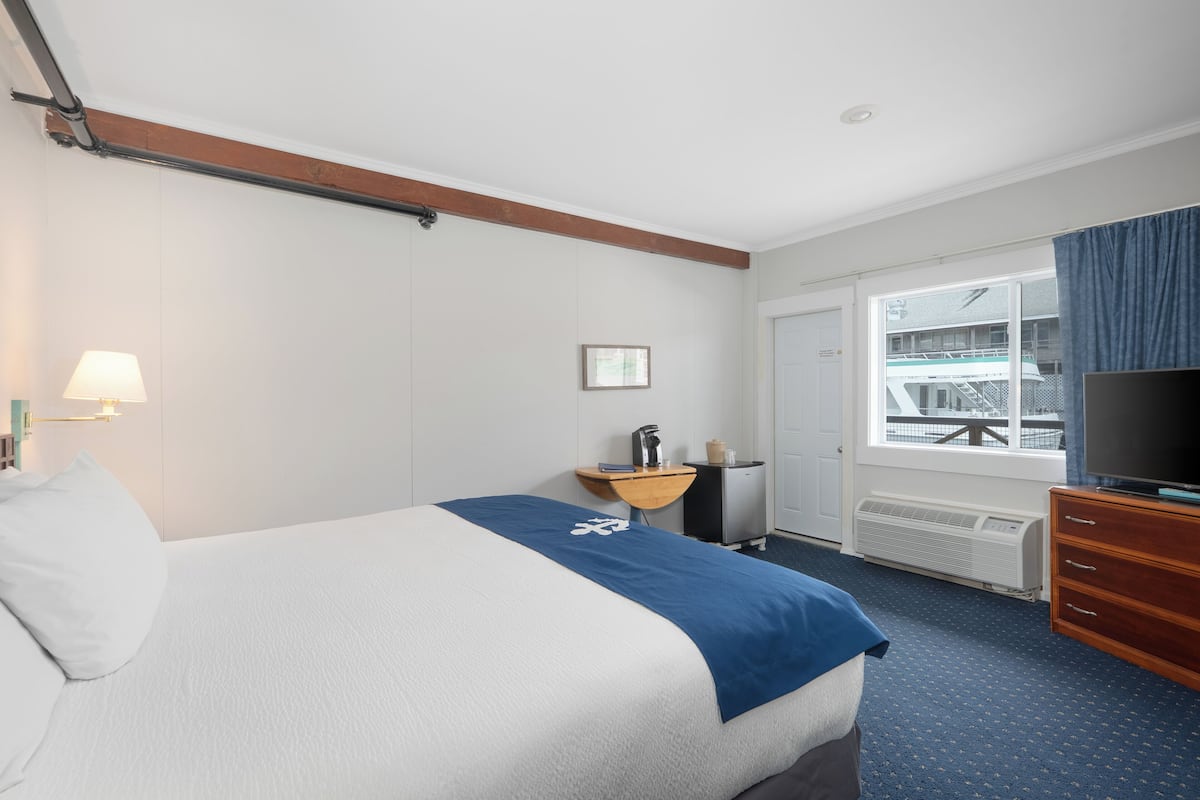 A hotel room with a bed, a TV, a window with a view of a building, a desk, and a lamp.