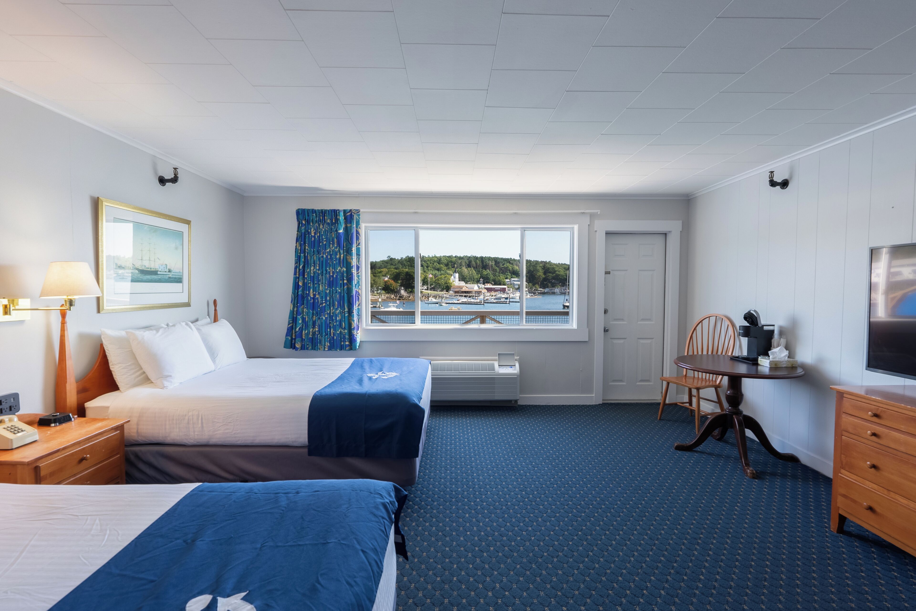 A hotel room with two beds, a large window with a view of a dock and boats, a desk with a chair, and a TV on the wall.