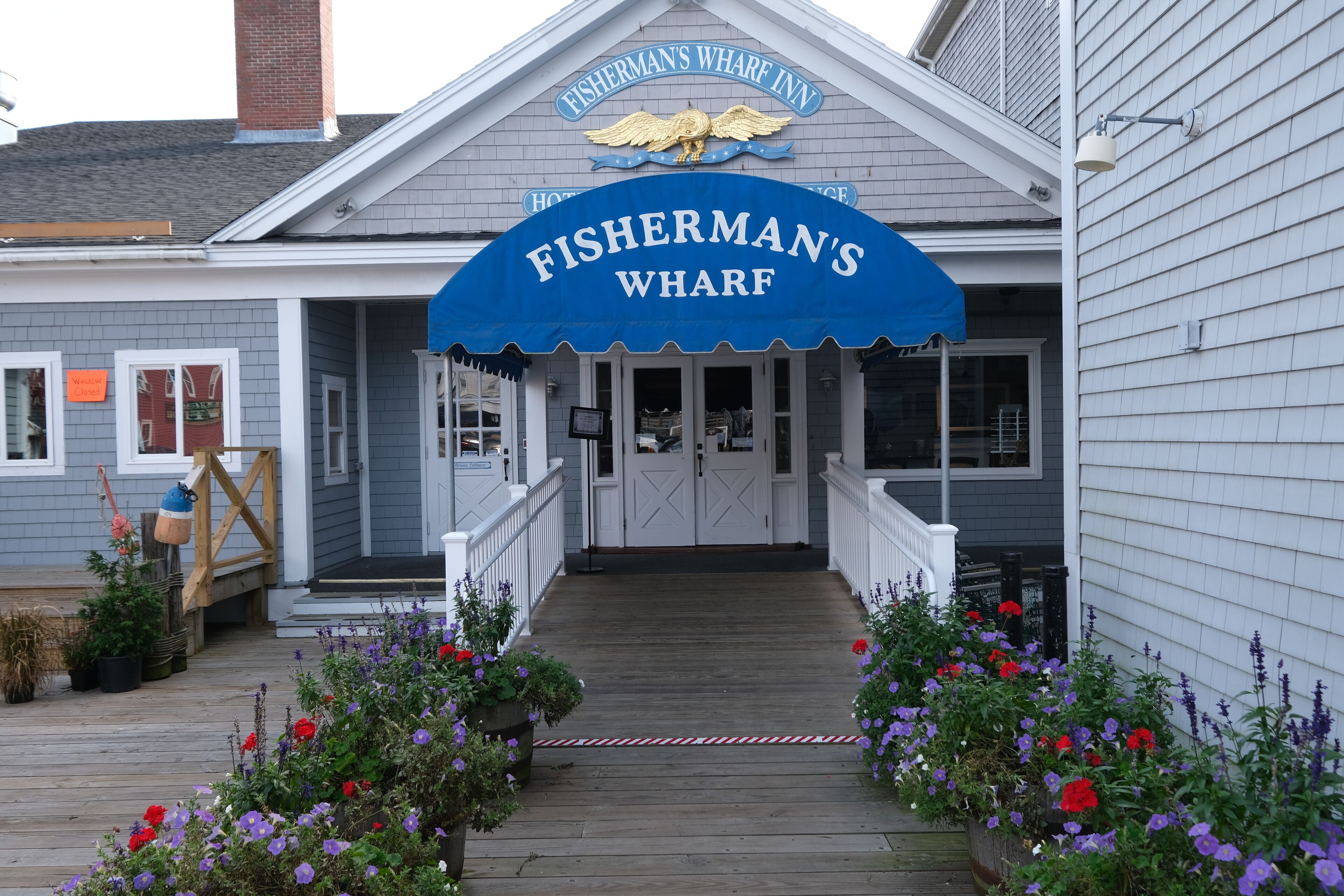 Das Hotel Fisherman's Wharf Inn mit blauem Markisenvorbau, Holzterrasse und Blumenbeeten.