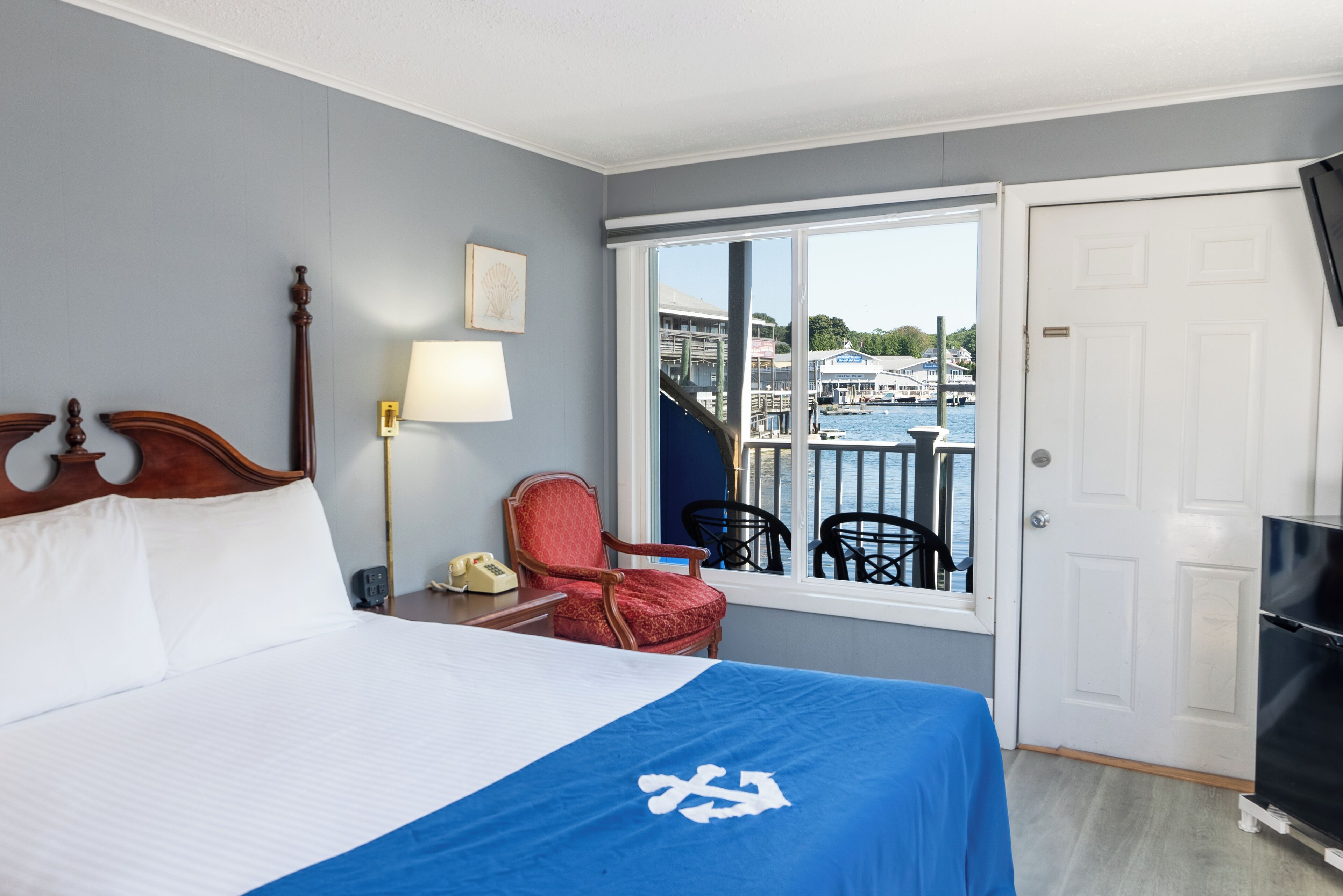 A hotel room with a large bed, a red chair, a desk with a lamp, and a view of a marina through a large window.