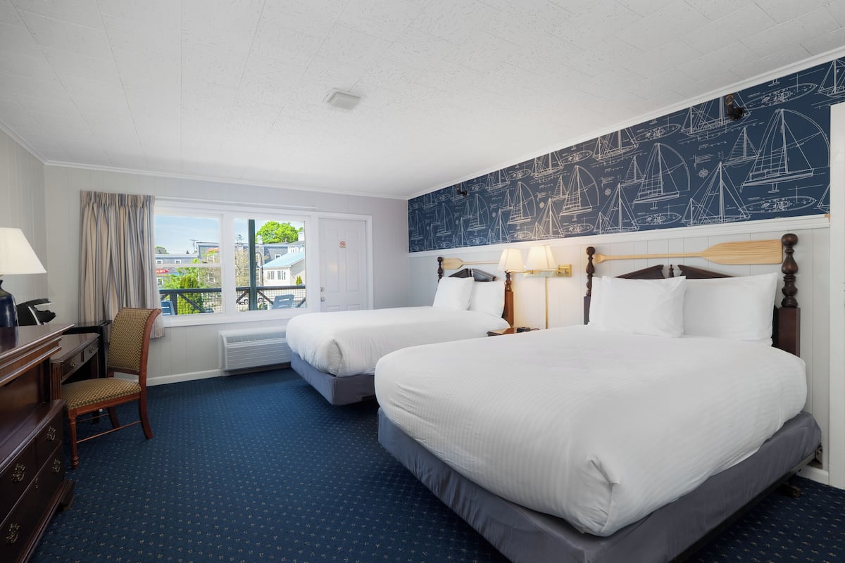 A hotel room with two beds, a desk, a chair, and a large window with a view of buildings and greenery.