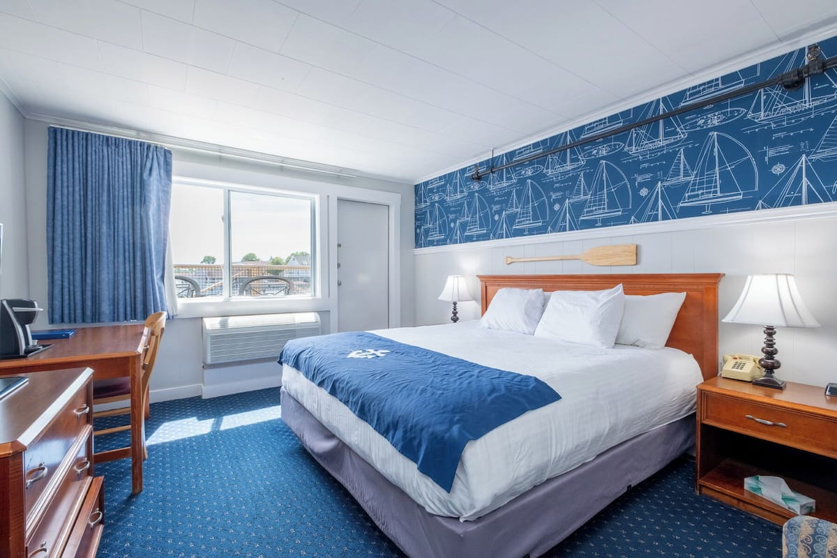 A hotel room with a large bed, a desk, a chair, a window with curtains, a wall with nautical-themed wallpaper, and a telephone on the nightstand.