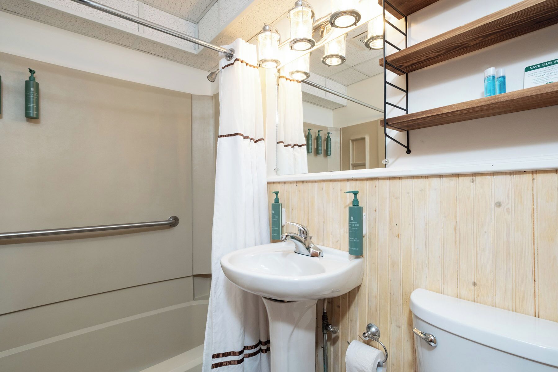 A bathroom with a white sink, a toilet, a shower with a white curtain, and a wooden shelf above the sink.