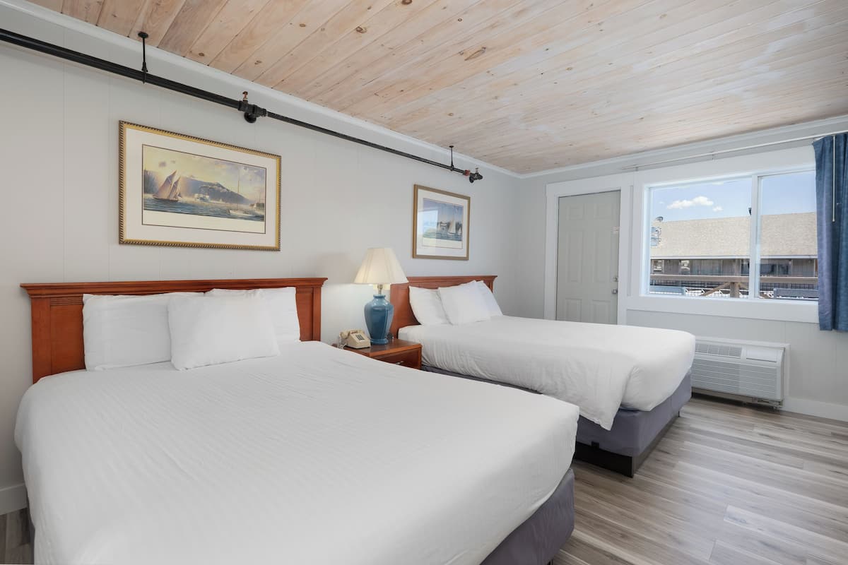 A hotel room with two beds, a wooden ceiling, framed pictures on the wall, a window with a view, and a nightstand with a lamp.