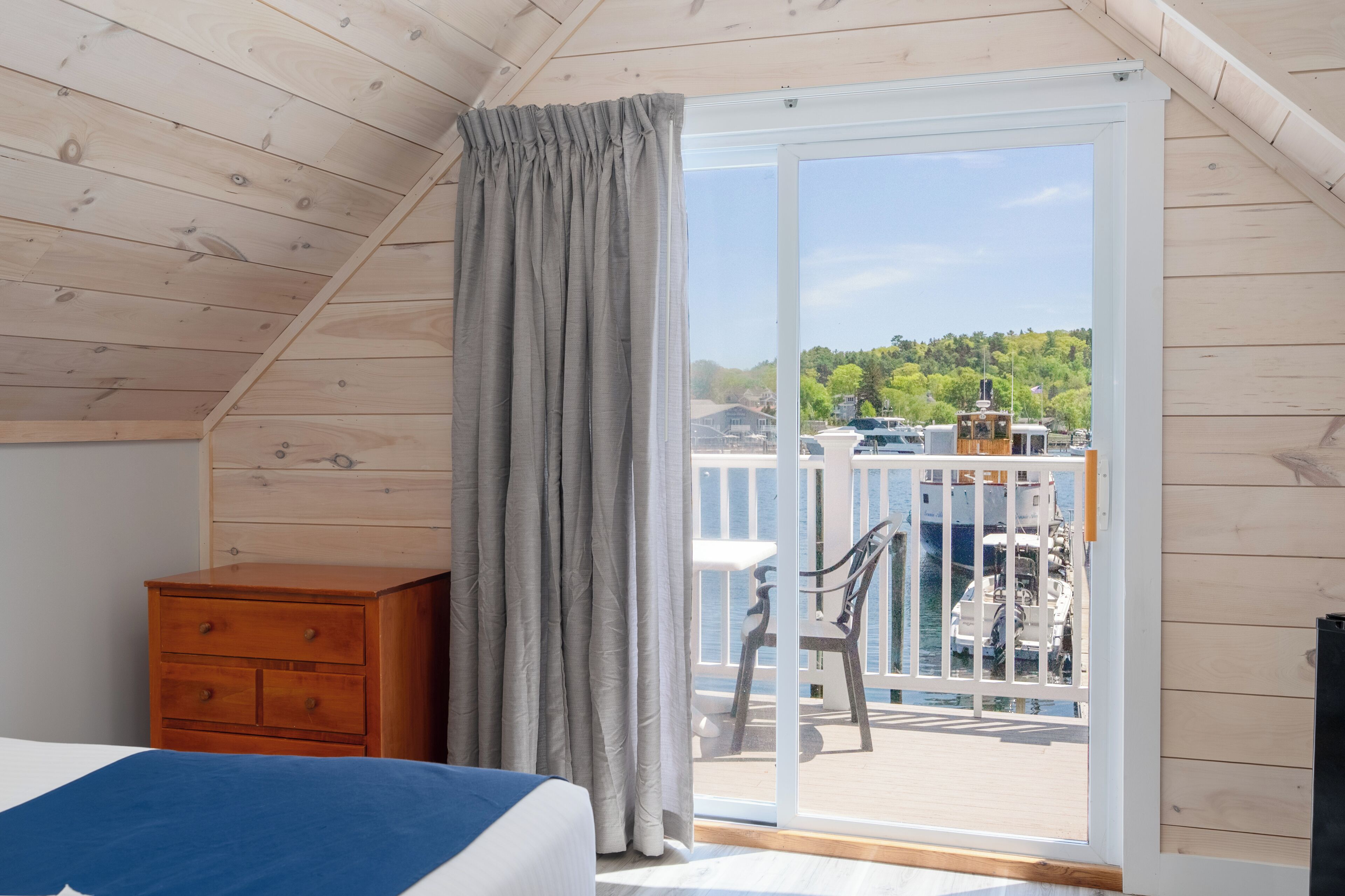 Una habitación con techo de madera, una cama, un aparador de madera y un ventanal corredero que da a una terraza exterior con vistas al mar y barcos.