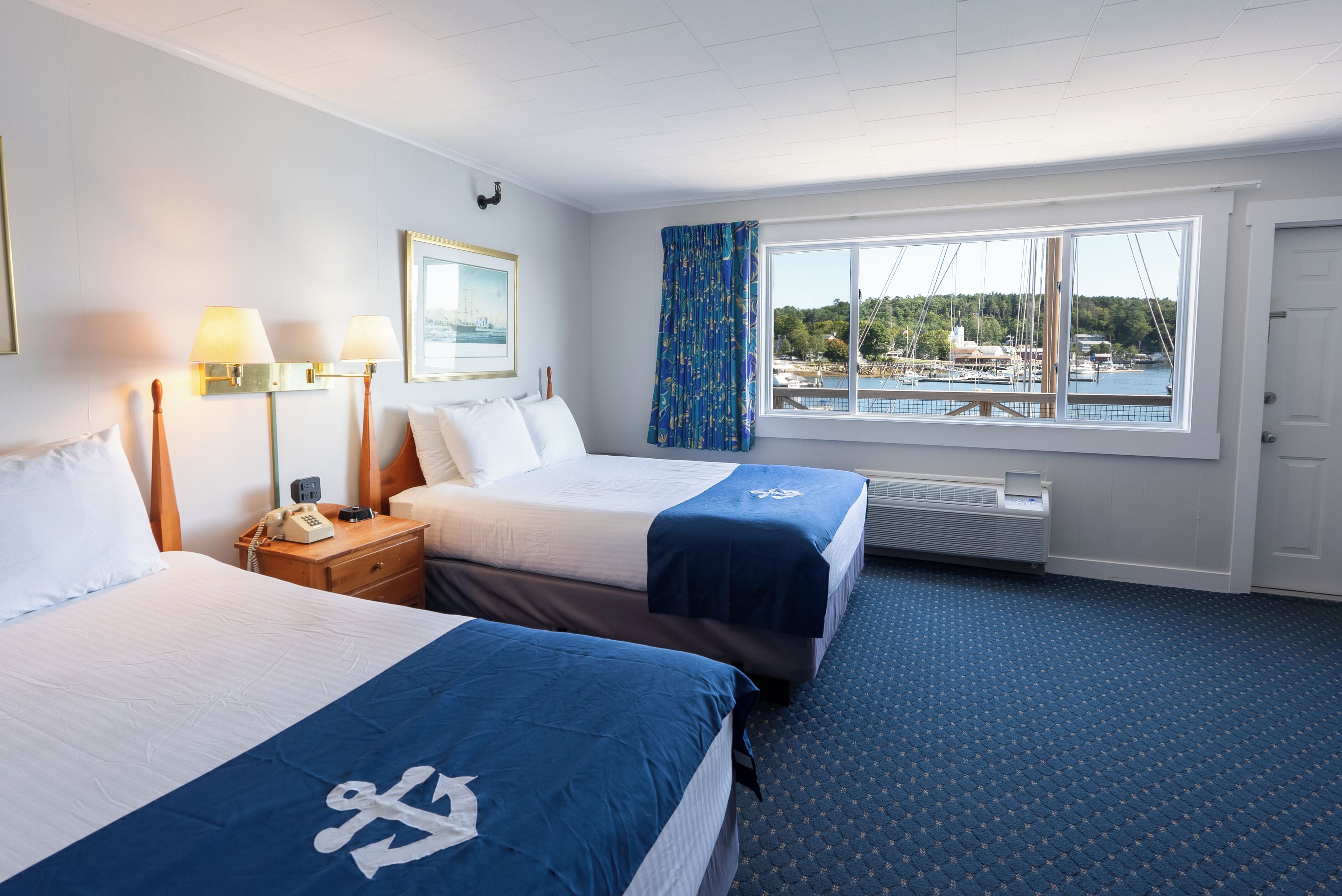 A hotel room with two beds, a large window with a view of a marina, a framed picture on the wall, and a telephone on the nightstand.
