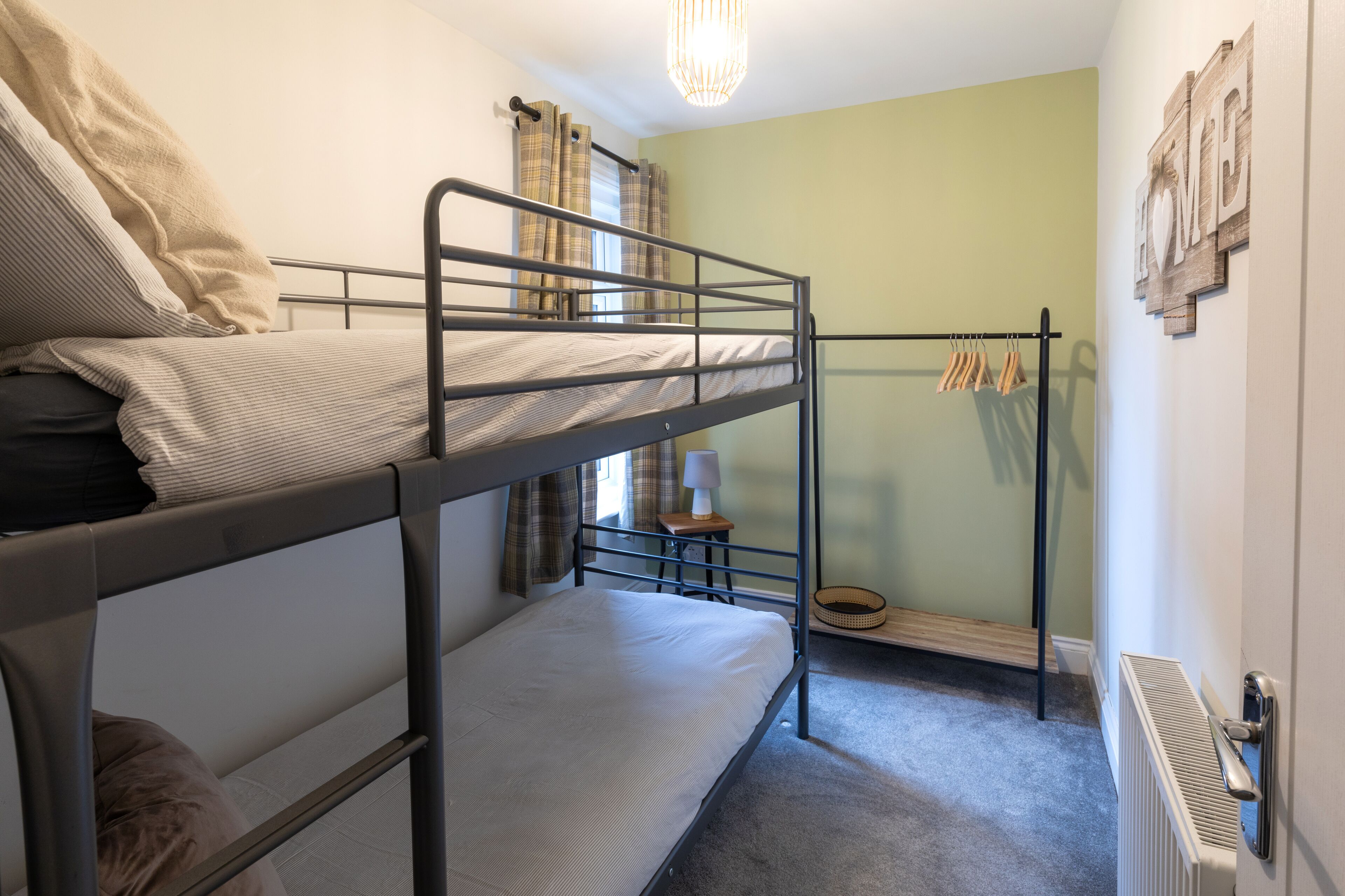 A bunk bed room with a radiator, a small table with a lamp, and a 'HOME' sign on the wall.