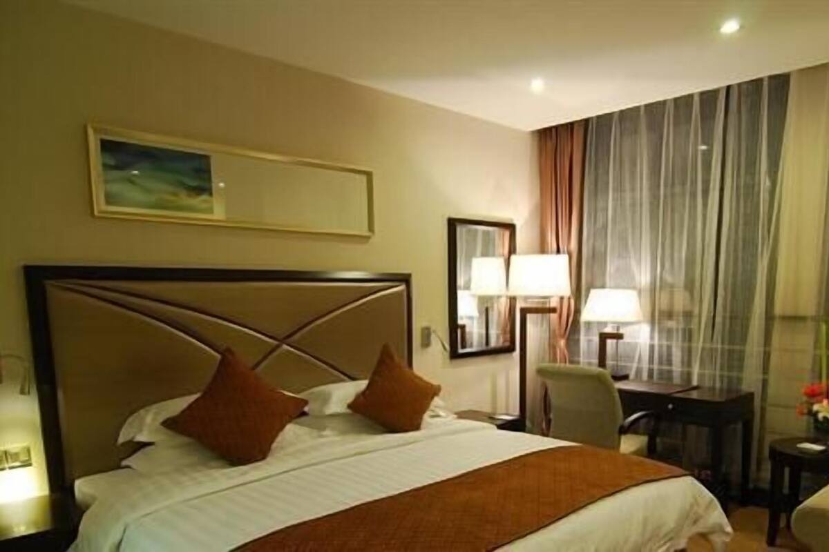 A hotel room with a large bed, a desk, a chair, and a view of the city through sheer curtains.