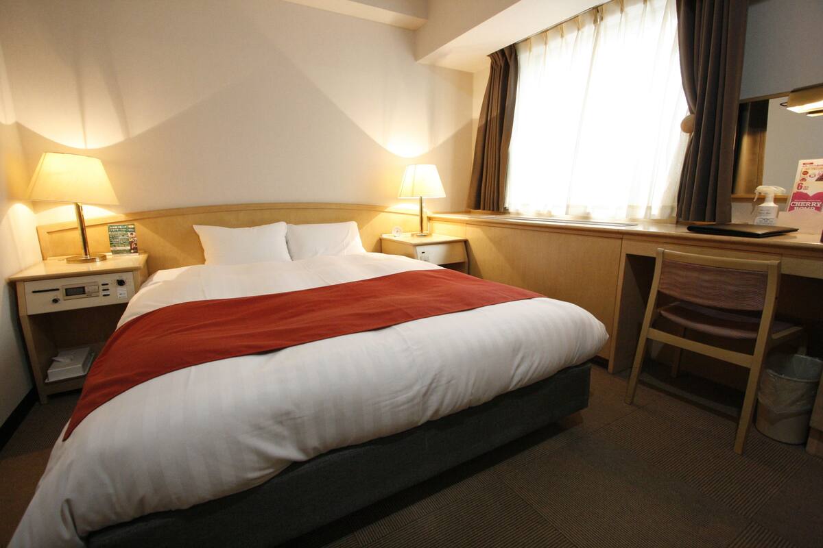 A hotel room with a bed, two bedside tables with lamps, a desk with a chair, and a window with curtains.