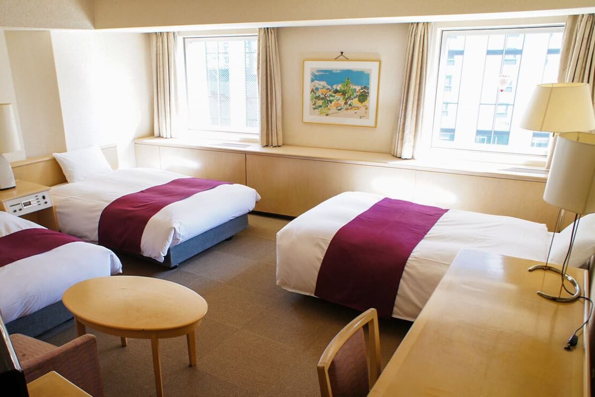 Hotellihuone, jossa on kaksi sänkyä, pieni pöytä, tuoli, työpöytä, lamppu, kello ja ikkuna, jossa on verhot.