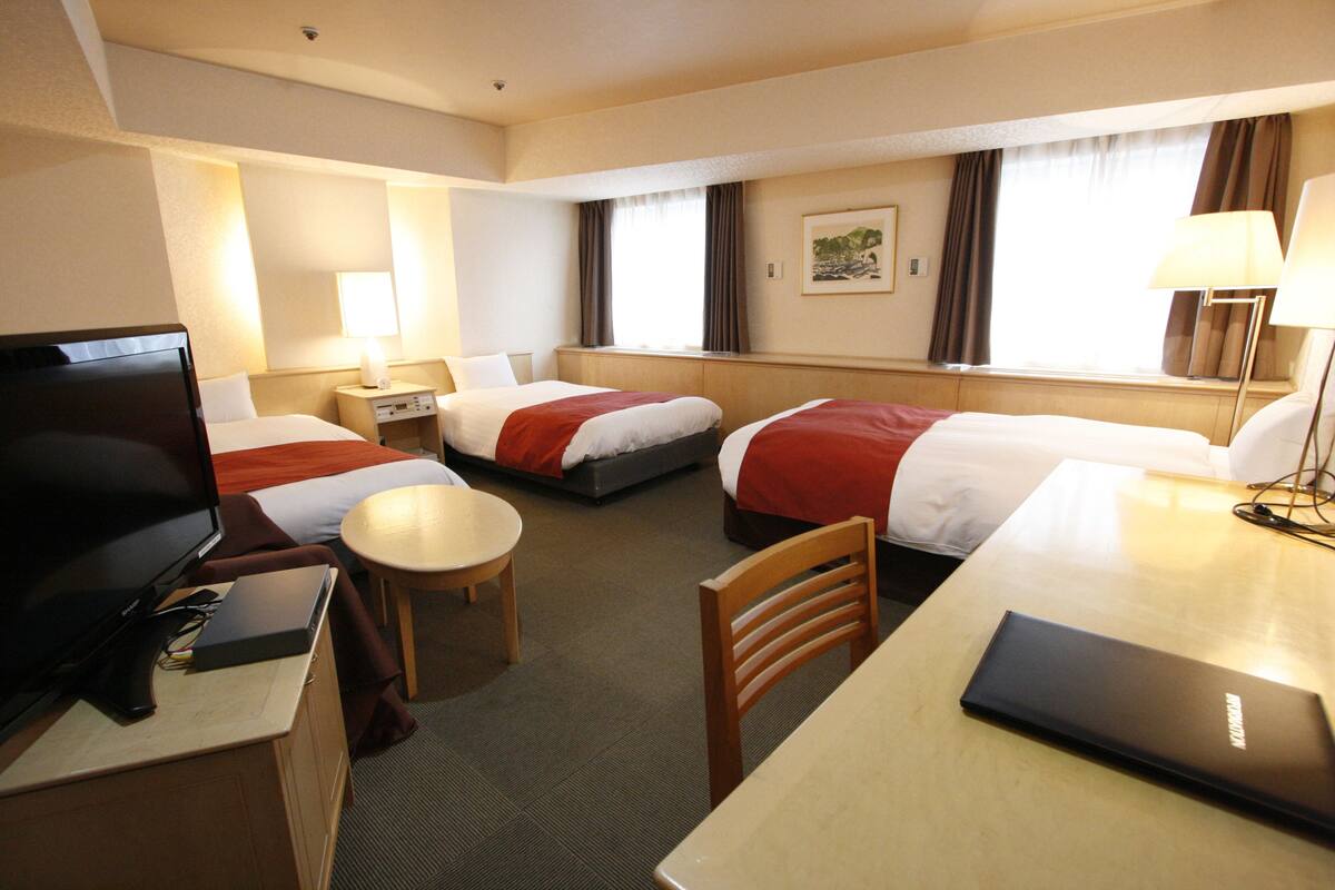 A hotel room with three beds, a desk, a chair, a television, and a lamp.