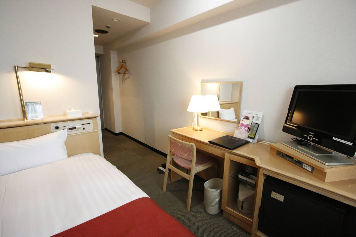 A hotel room with a bed, desk, chair, TV, and a trash bin.