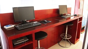 Un escritorio rojo con dos monitores, un teclado y un ratón. Hay dos taburetes negros y un estante pequeño con un dispositivo.