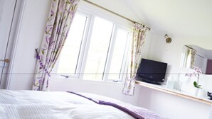 A bedroom with a bed, a TV on a shelf, a window with curtains, and a small plant. - Quixhill - Denstone - Dove Meadow B & B (Uttoxeter)