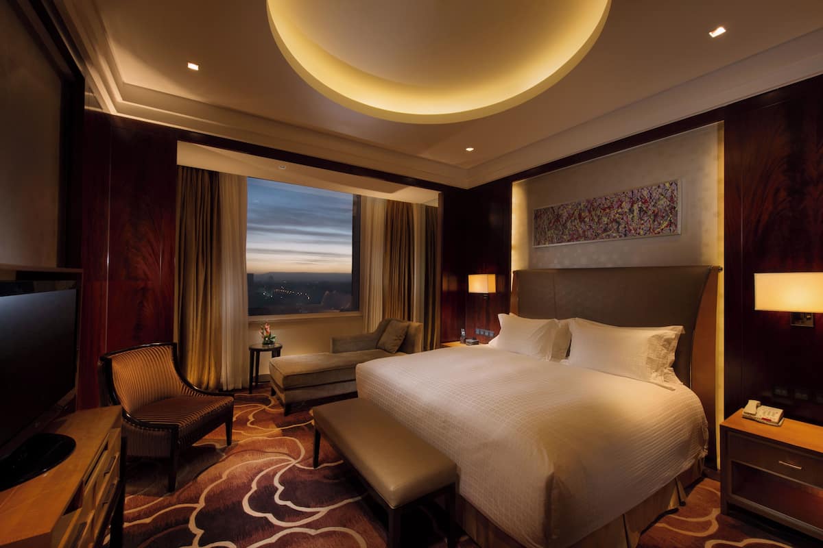A hotel room with a large bed, a desk with a chair, a small table, and a window with curtains.