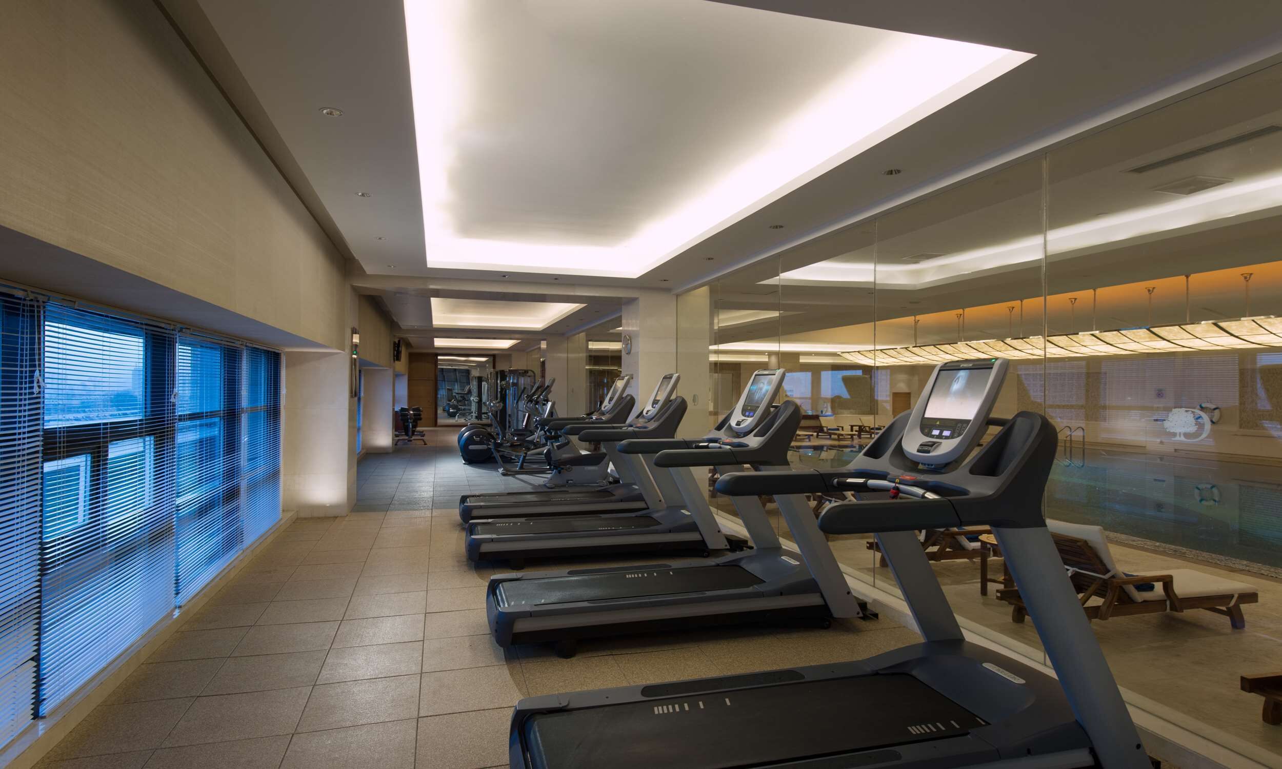 A fitness center with treadmills aligned along a corridor, large windows on one side, and a mirrored wall on the other.