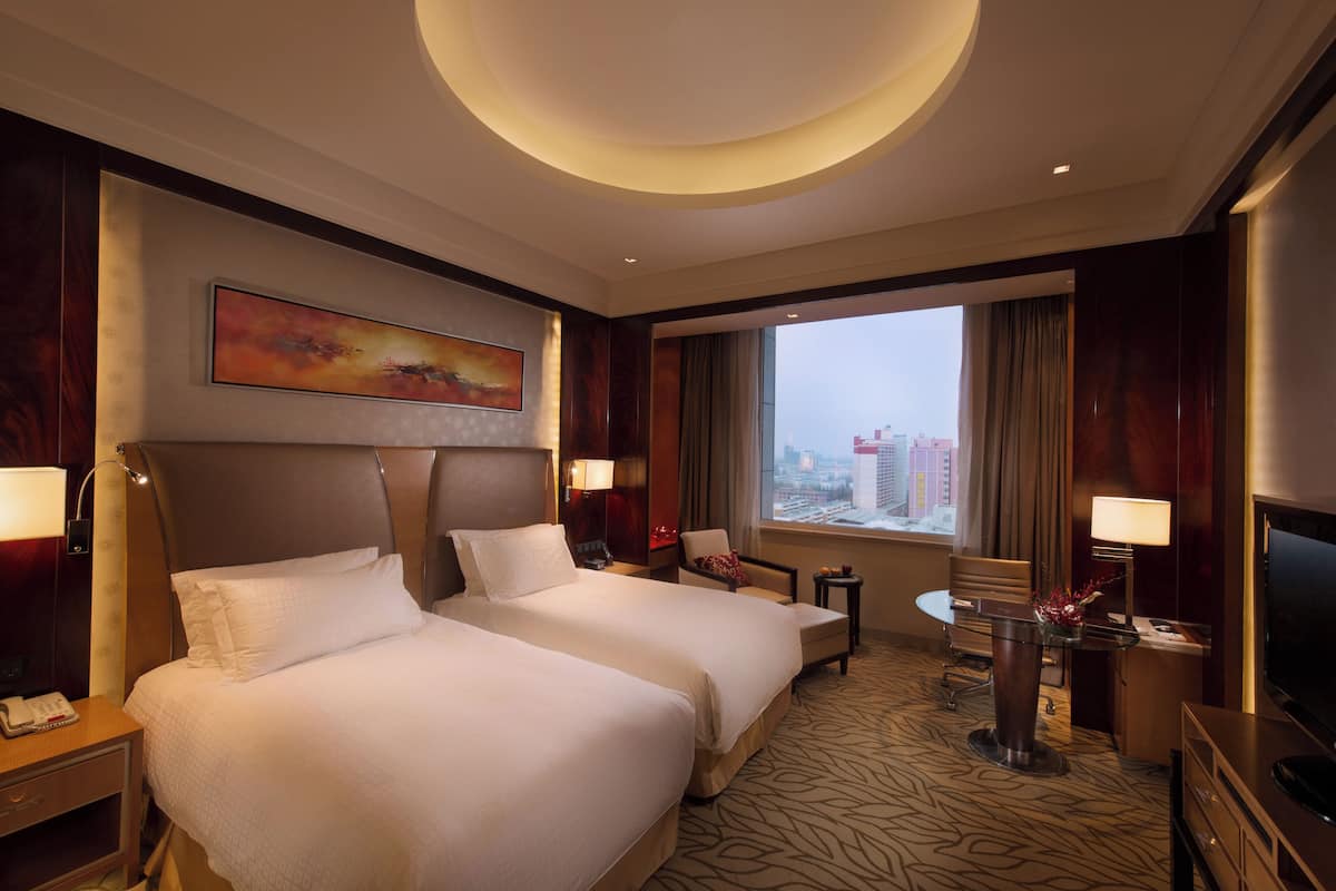 A hotel room with two beds, a desk, a chair, a TV, and a large window with a city view.