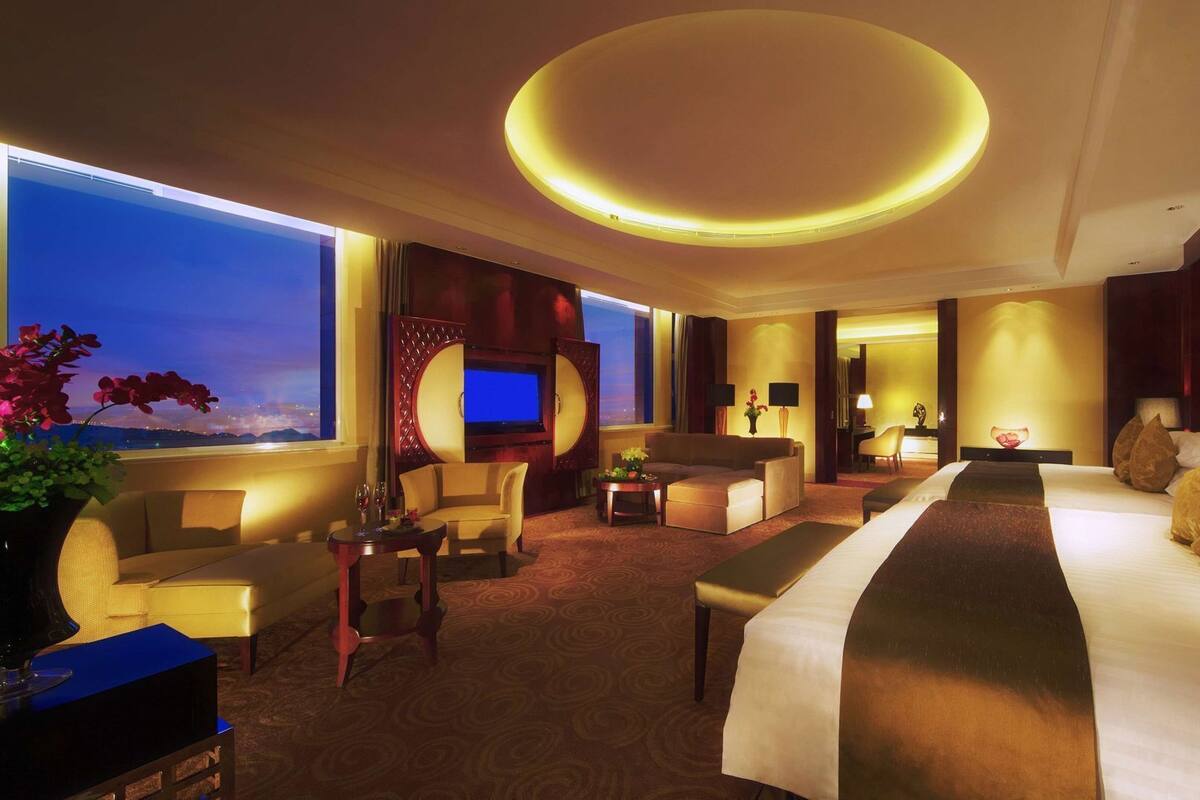A hotel room with a large bed, a sofa, a TV, and a view of the city at dusk.