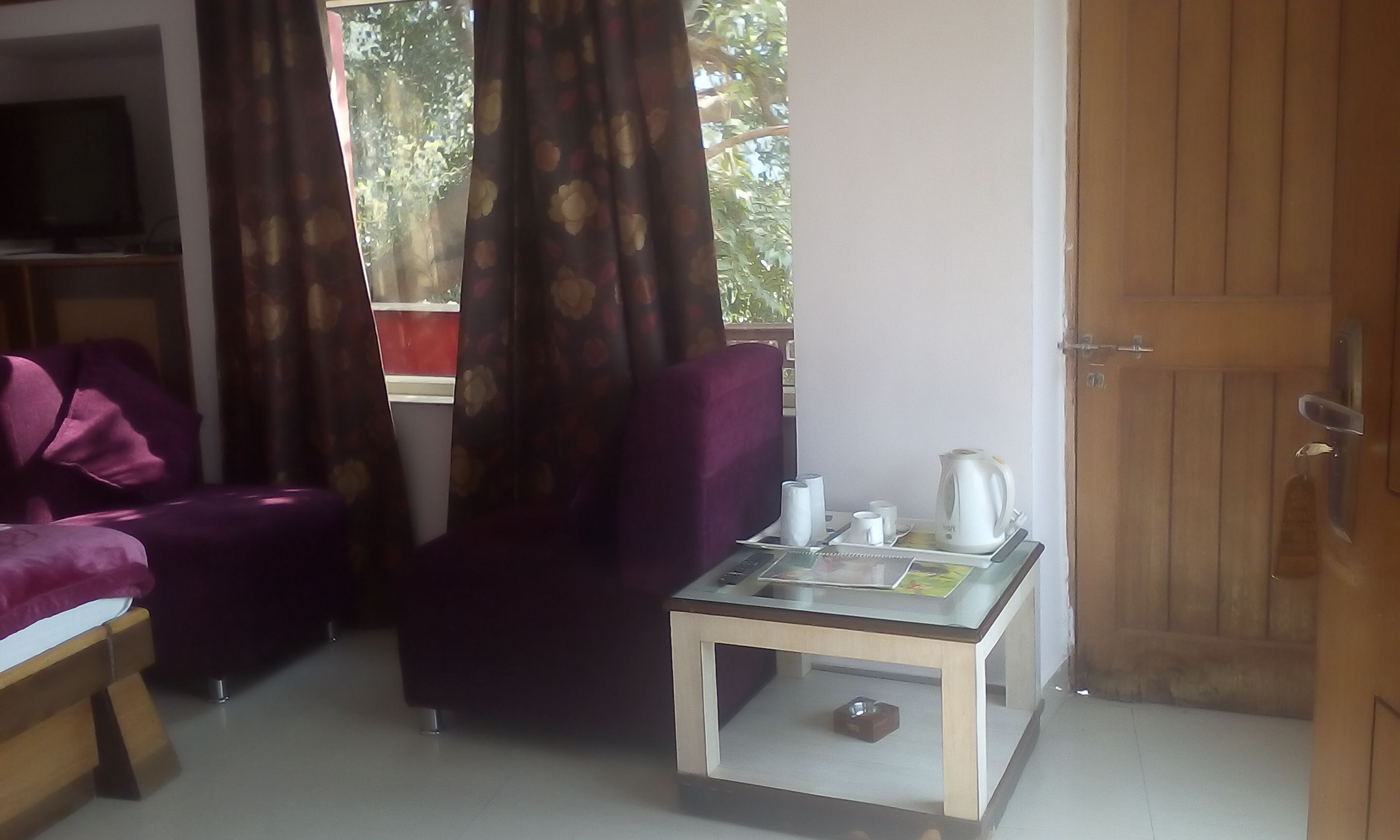 Un salón con un sofá púrpura, una mesa de centro de cristal con una tetera, tazas y un libro, y una puerta de madera.