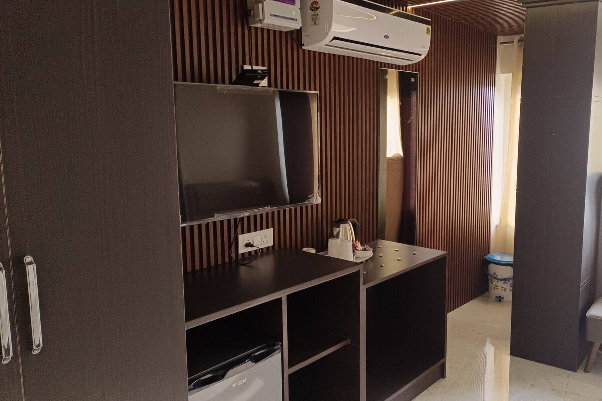 Habitación de hotel con televisión de pantalla plana, cafetera, basurero y zona de estar con una silla y reposapiés.