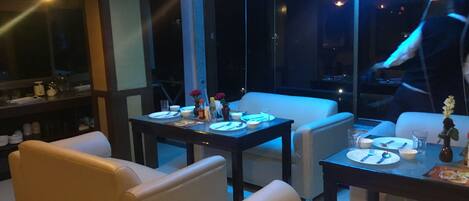 A modern dining area with a table set for two, a sofa, and a large window with a view of the city at night.