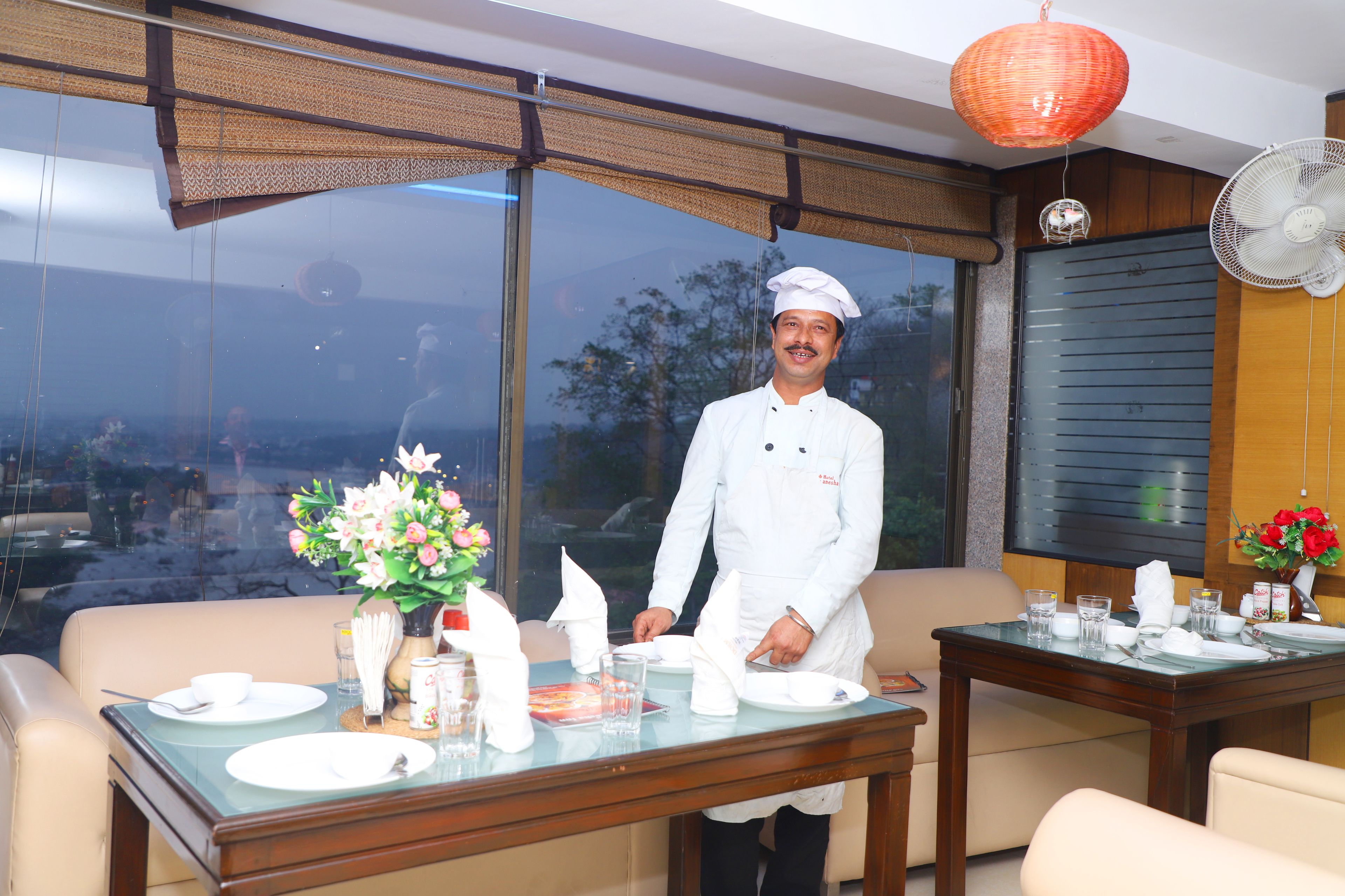 A dining area with a table set for two, a chef in uniform, and a large window with a view.