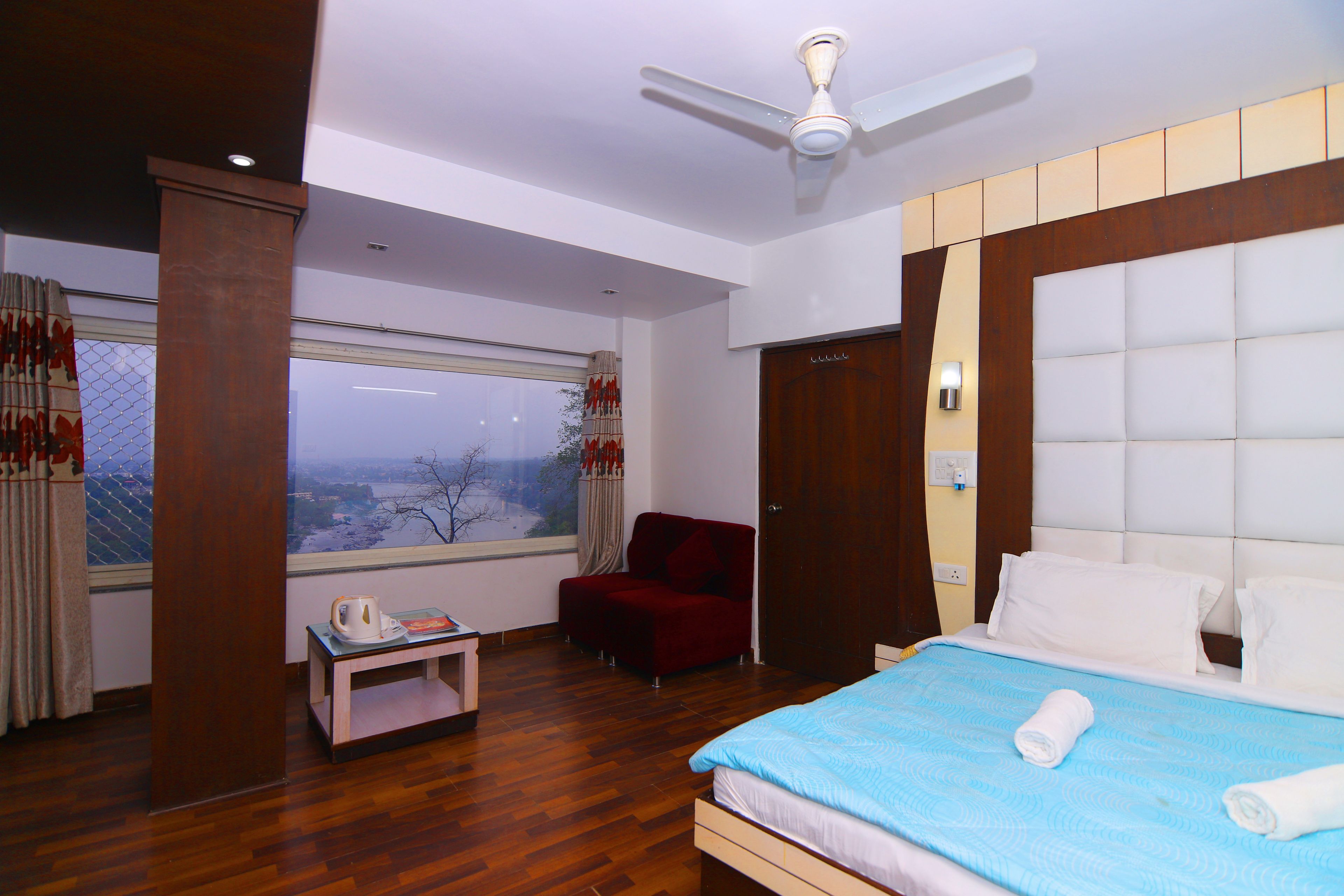 A hotel room with a large bed, a ceiling fan, a wooden floor, a small table with a kettle, a red chair, and a window with a view.