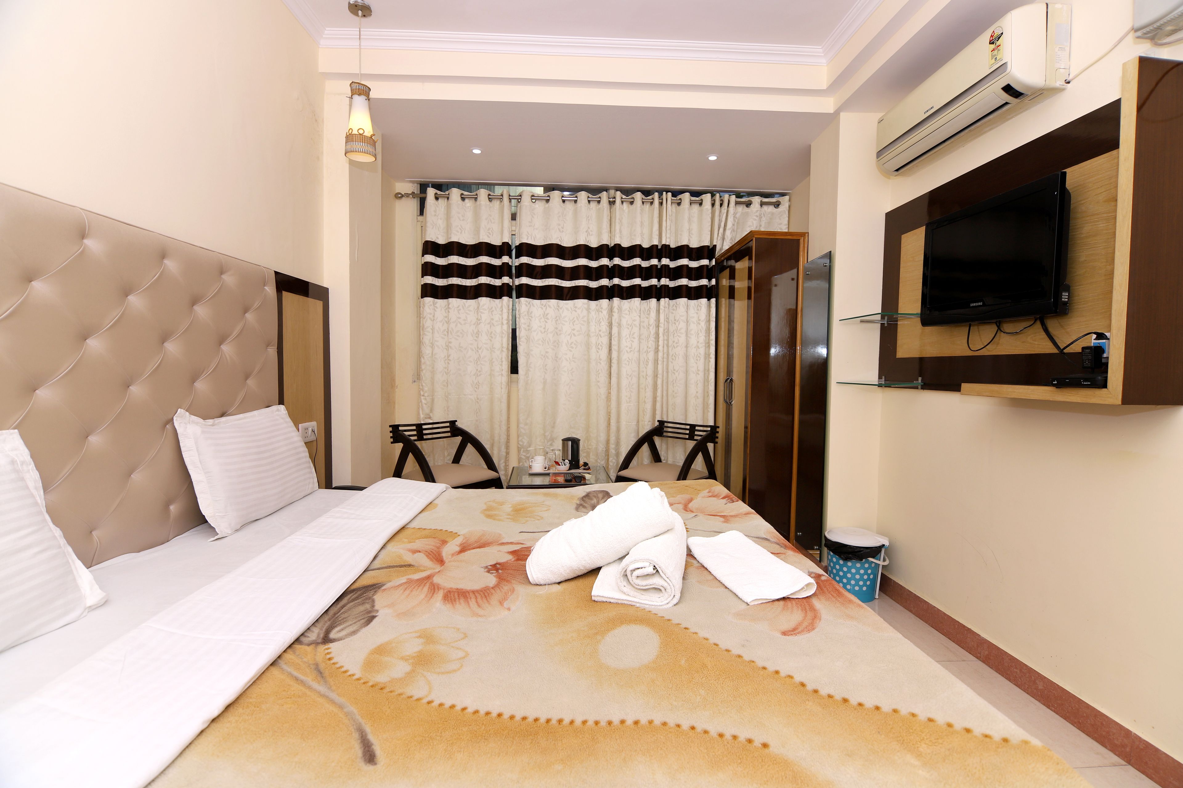 A hotel room with a large bed, a television mounted on the wall, a small table with chairs, and a window with curtains.