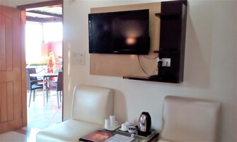 A room with a flat-screen TV mounted on the wall, two white chairs, a small table with a kettle, and a window overlooking an outdoor area with chairs and a table.