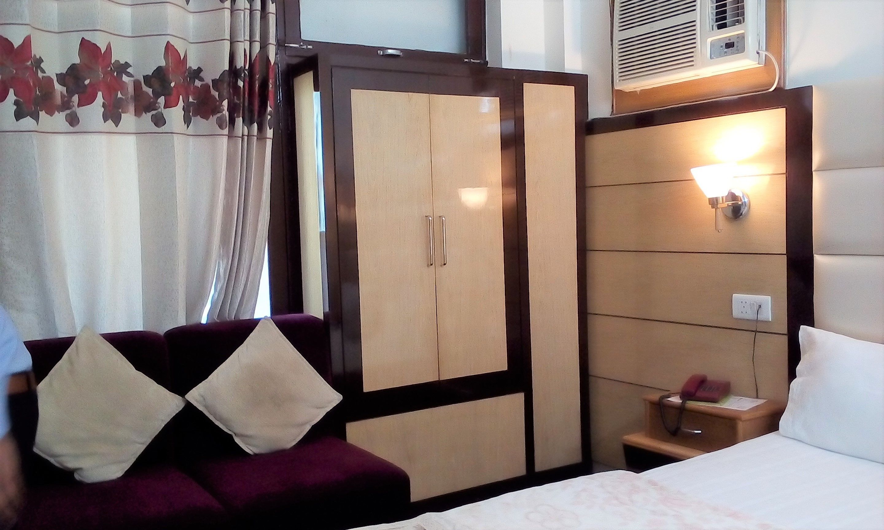 A hotel room with a bed, a nightstand, a wardrobe, and a seating area with a sofa and pillows.