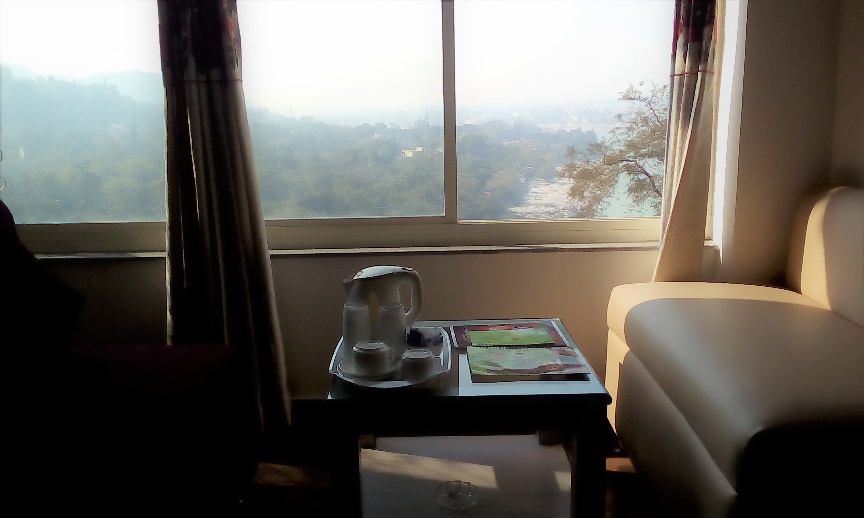 A room with a view of a mountainous landscape through a large window. There is a small table with a kettle, cups, and a magazine. A chair is positioned near the window.