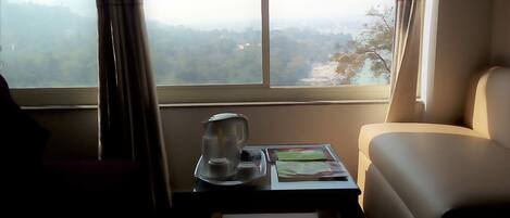 A room with a view of a mountainous landscape through a large window. There is a small table with a kettle, cups, and a magazine. A chair is positioned near the window.