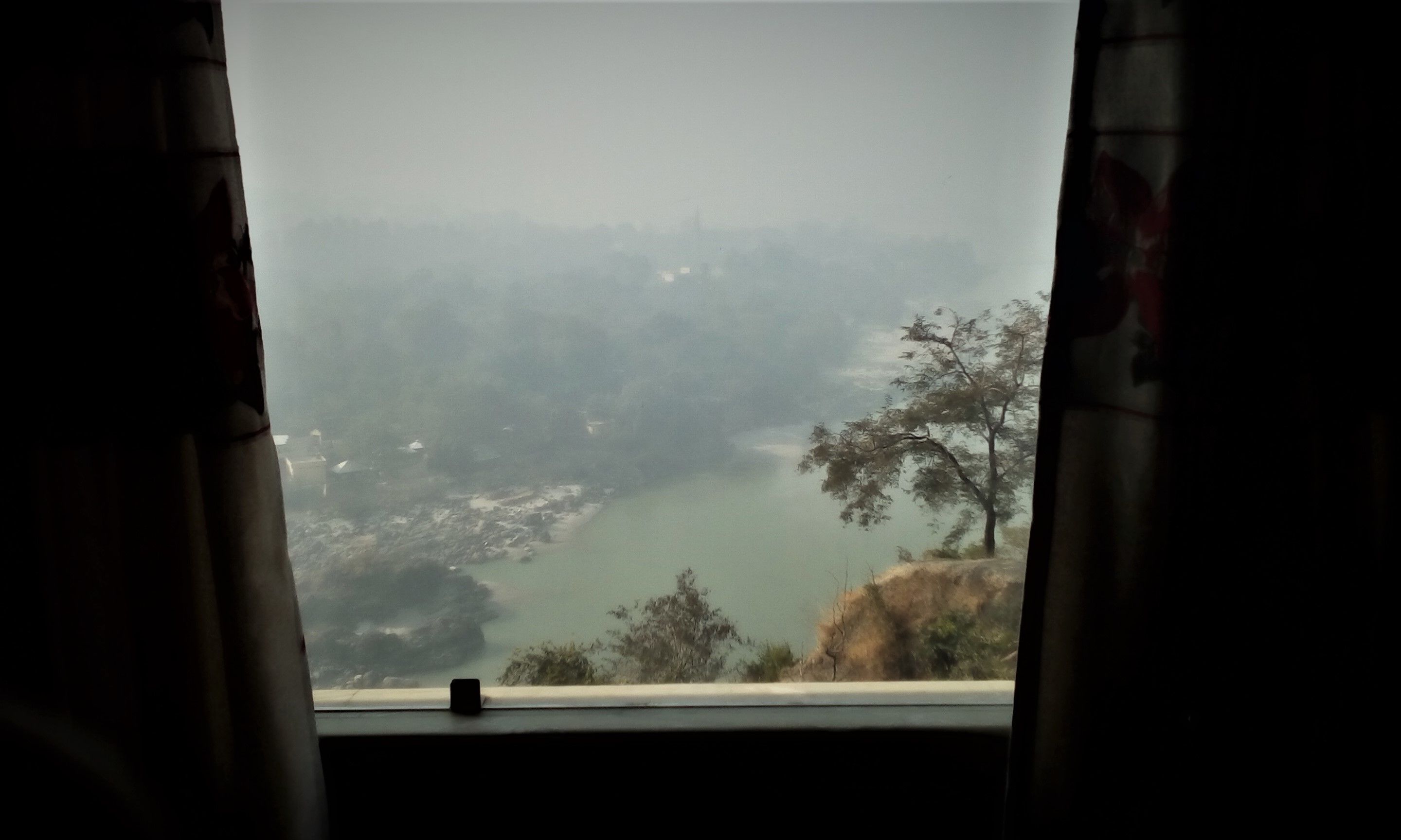 A view from a window with curtains, overlooking a valley with trees and a body of water.