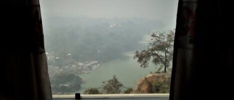 A view from a window with curtains, overlooking a valley with trees and a body of water.