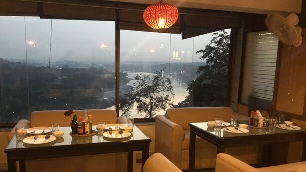Uma área de refeições com uma mesa posta para dois, um vaso com uma única flor, e vista para a cidade e rio através de grandes janelas.