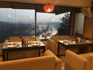 Un comedor con una mesa preparada para dos, un jarrón con una sola flor, y vistas a un paisaje urbano y un río a través de grandes ventanales.