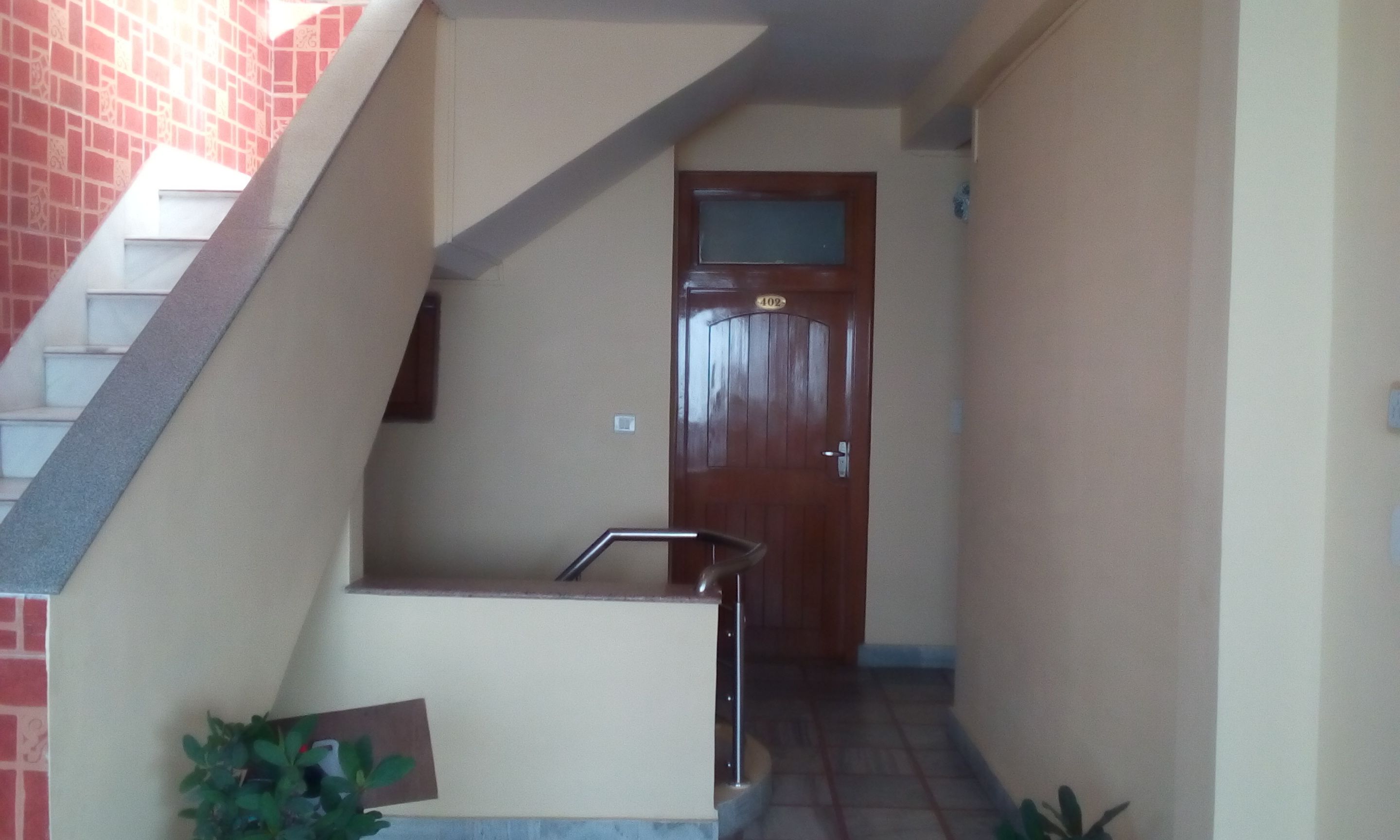 A narrow hallway with a wooden door marked "402" and a small outdoor area with a bench and potted plants.